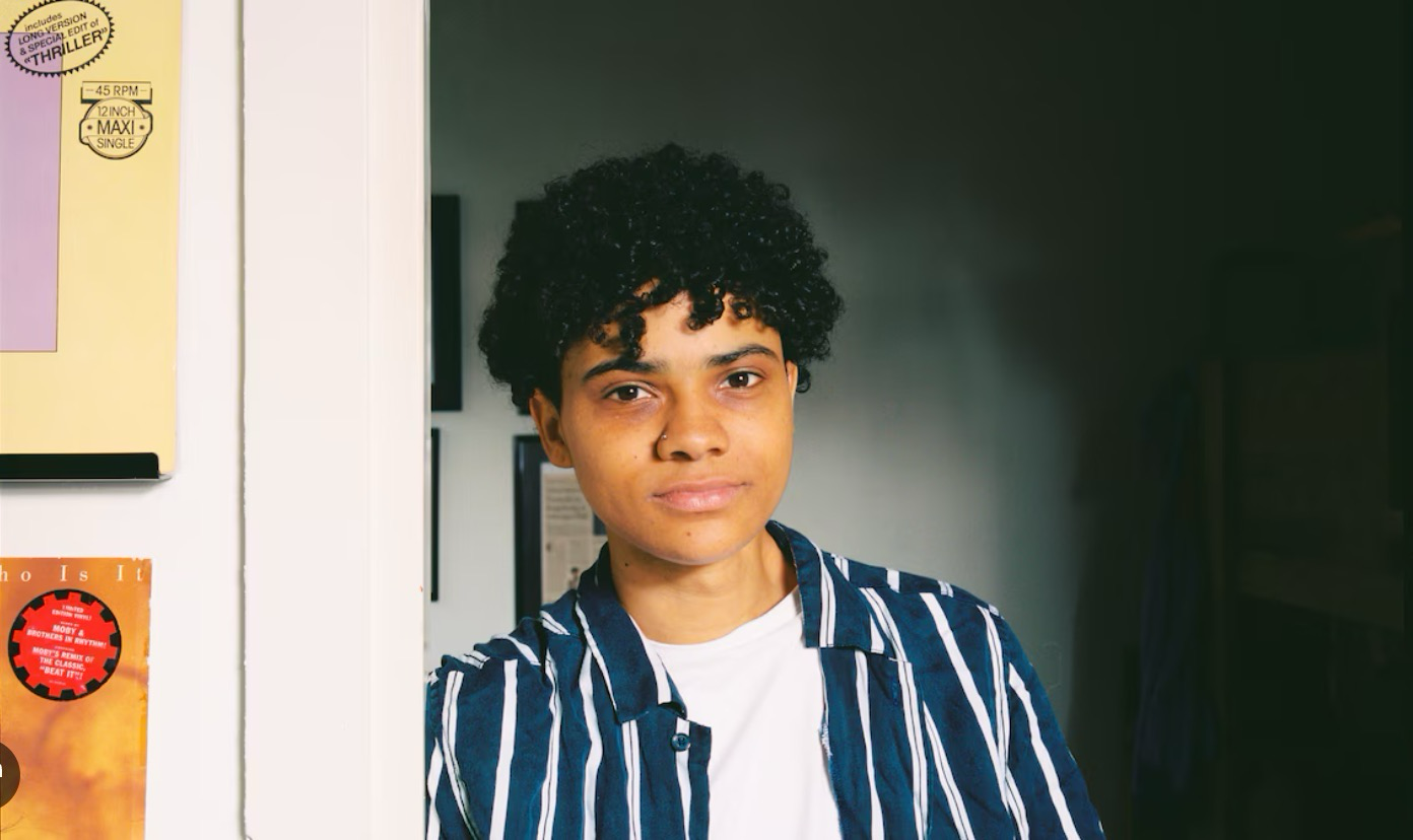 Portret van Jason Bhugwandass, staand in de deuropening van zijn kamer.
                  Achter hem hangen tekeningen en platenhoezen.